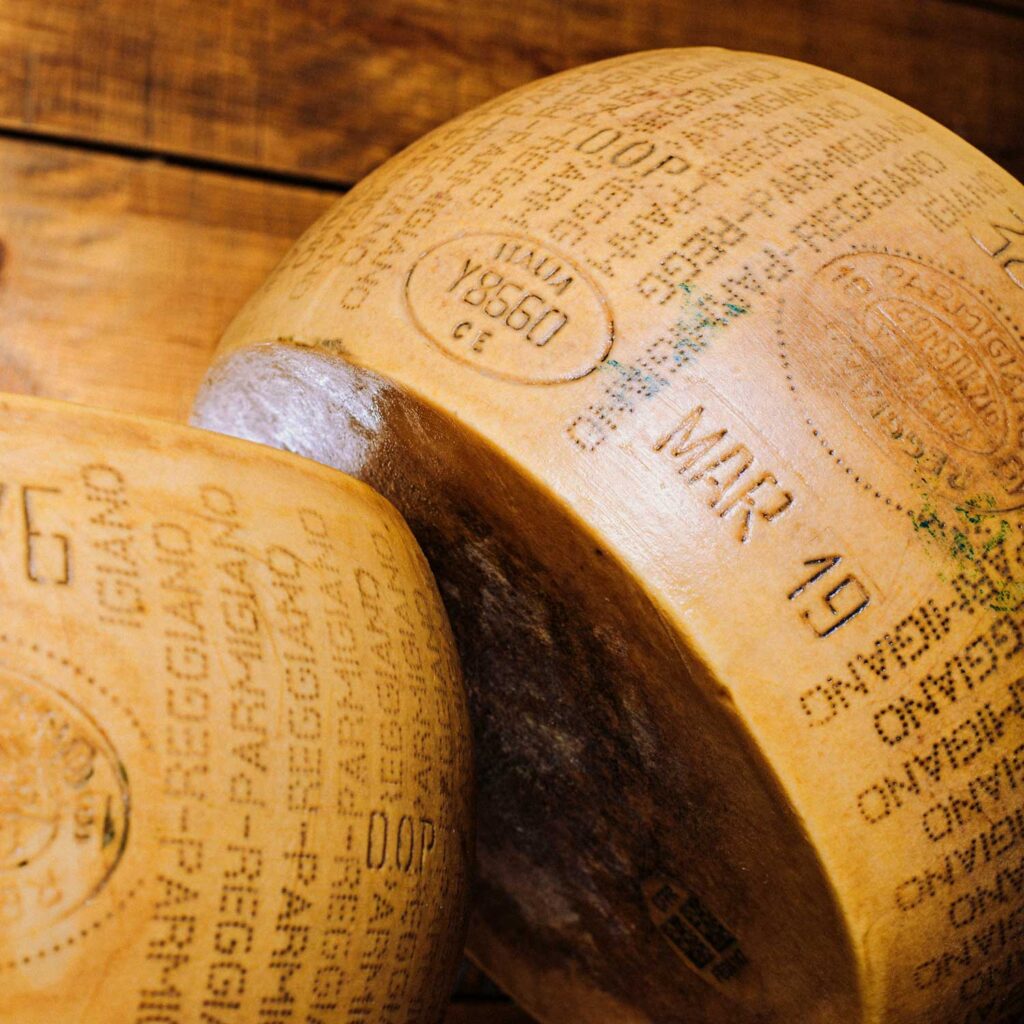 Nahaufnahme zweier großer Parmigiano Reggiano-Käseräder, die auf einer rustikalen Holzfläche liegen. Die Prägungen mit dem Herkunftsnachweis und das raue, kristallisierte Innere des Käses sind deutlich sichtbar und zeigen die hohe Qualität dieses traditionellen italienischen Produkts.