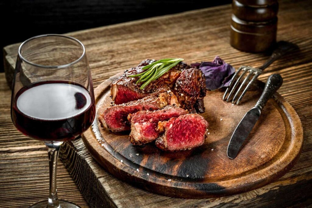 Ein Glas tiefroter Primitivo-Wein neben einem saftigen, perfekt gegrillten Steak auf einem rustikalen Holzbrett – die perfekte Kombination für Fleischliebhaber.