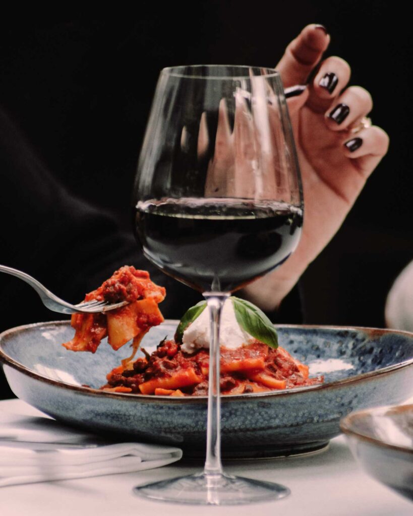 Eine Frau hebt eine Gabel mit frischer Pasta in würziger Tomatensauce, während ein Glas Primitivo-Wein daneben auf dem Tisch steht – ein echter Genussmoment der italienischen Küche.