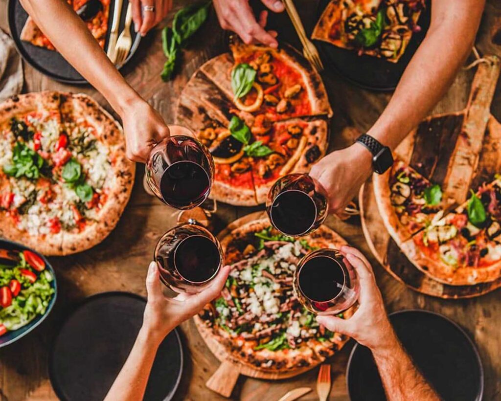 Freunde stoßen mit Gläsern Primitivo-Wein an, während verschiedene Pizzen mit frischen Zutaten auf dem Tisch stehen – italienische Lebensfreude pur!