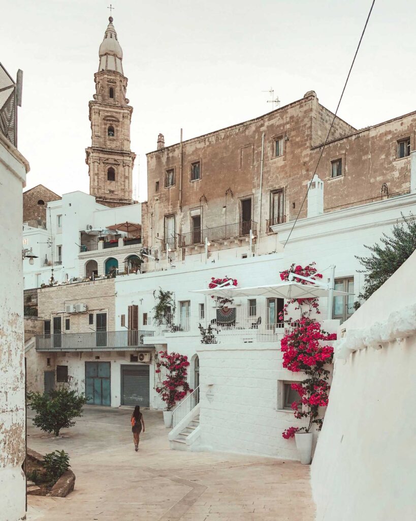 Eine malerische Gasse in einer historischen Stadt Apuliens, gesäumt von weißen Häusern mit blühenden Bougainvillea. Im Hintergrund ragt ein beeindruckender Kirchturm in den Himmel.