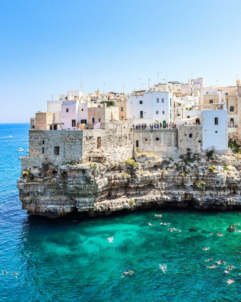 Die Küstenstadt Polignano a Mare thront auf majestätischen Kalksteinklippen über dem tiefblauen Meer. Menschen schwimmen im klaren Wasser unterhalb der weißen Häuser.