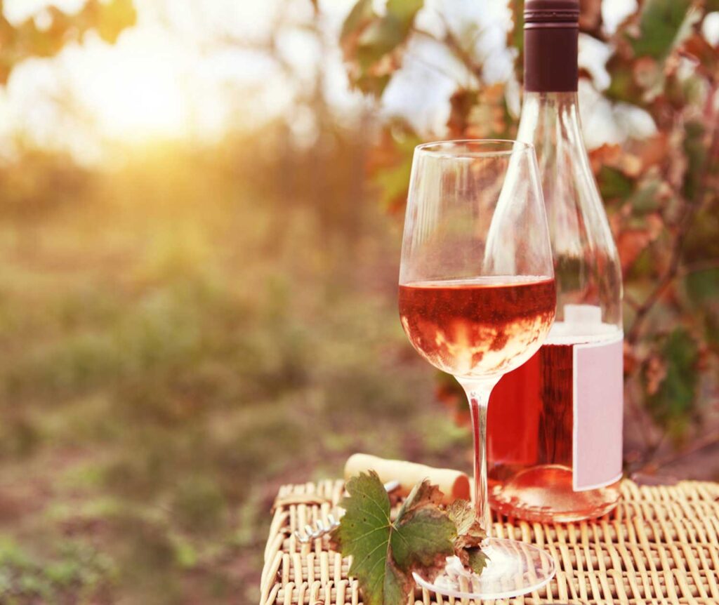 Ein Glas leuchtend roséfarbener Primitivo Wein steht auf einem geflochtenen Tisch im Freien, daneben eine geöffnete Flasche. Im Hintergrund verschwimmen herbstliche Weinreben und die warme Abendsonne taucht die Szene in goldenes Licht – ein perfekter Moment zum Genießen.