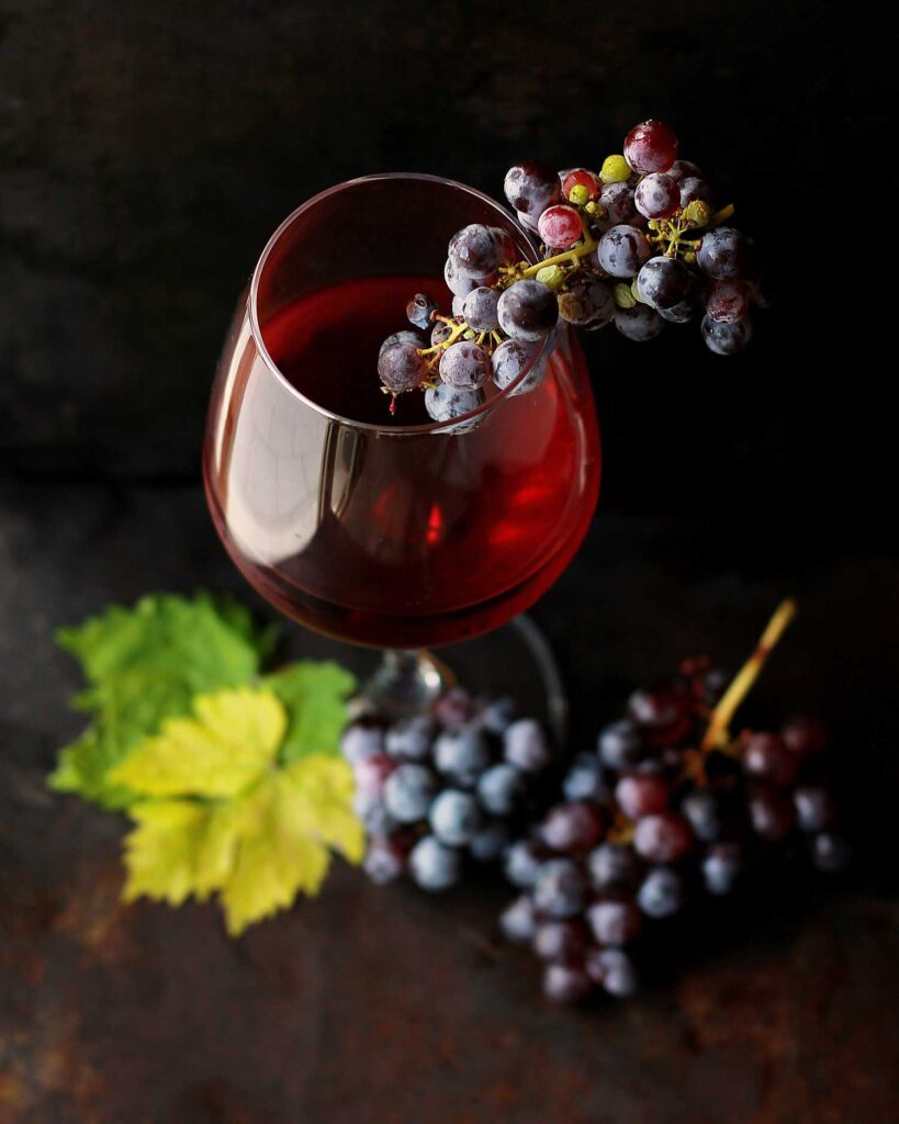 Ein Glas tiefroter Lambrusco-Wein mit einer kleinen, reifen Weintraube, die über den Rand gelegt ist. Im Hintergrund liegen weitere dunkle Trauben und ein frisches, grünes Weinblatt auf einer rustikalen, dunklen Oberfläche.