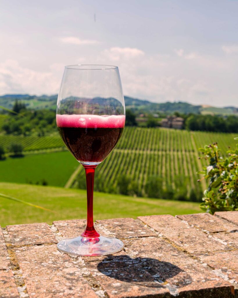 Ein Glas sprudelnder Lambrusco auf einer Steinmauer mit Blick auf grüne Weinberge in der Emilia-Romagna unter blauem Himmel.