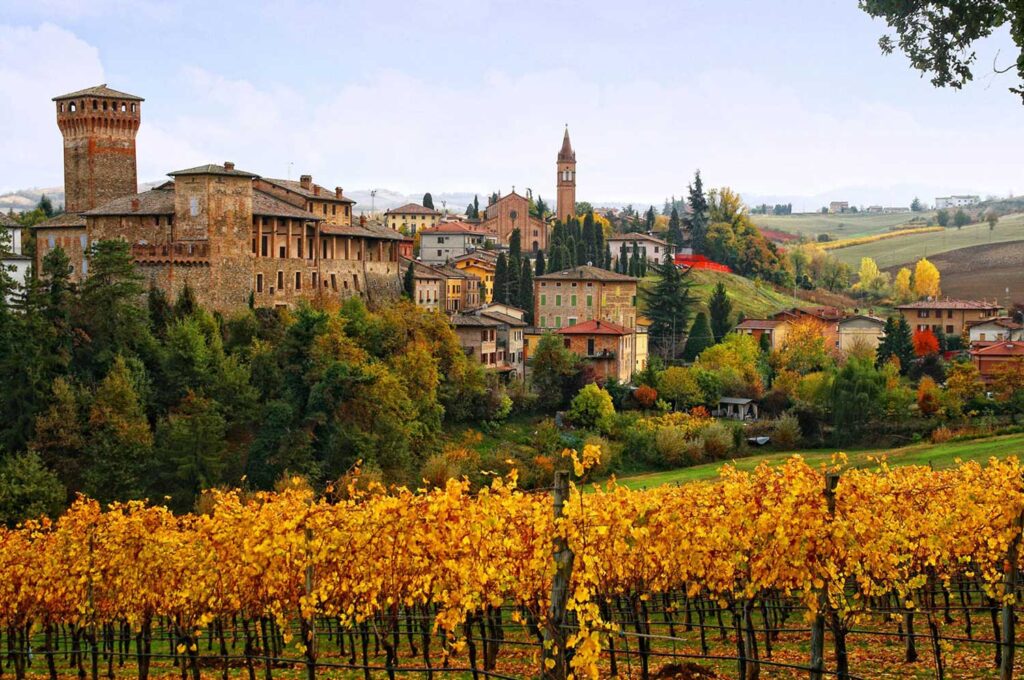 Eine malerische italienische Landschaft mit einer Burg, einem Dorf und herbstlichen Lambrusco Weinbergen in warmen Farben.