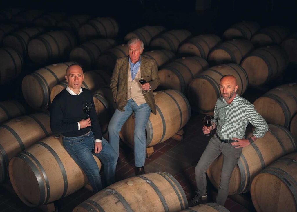 Drei Männer sitzen auf großen Weinfässern in einem dunklen Lambrusco Weinkeller, halten Gläser mit Lambrusco Rotwein in der Hand und blicken in die Kamera.