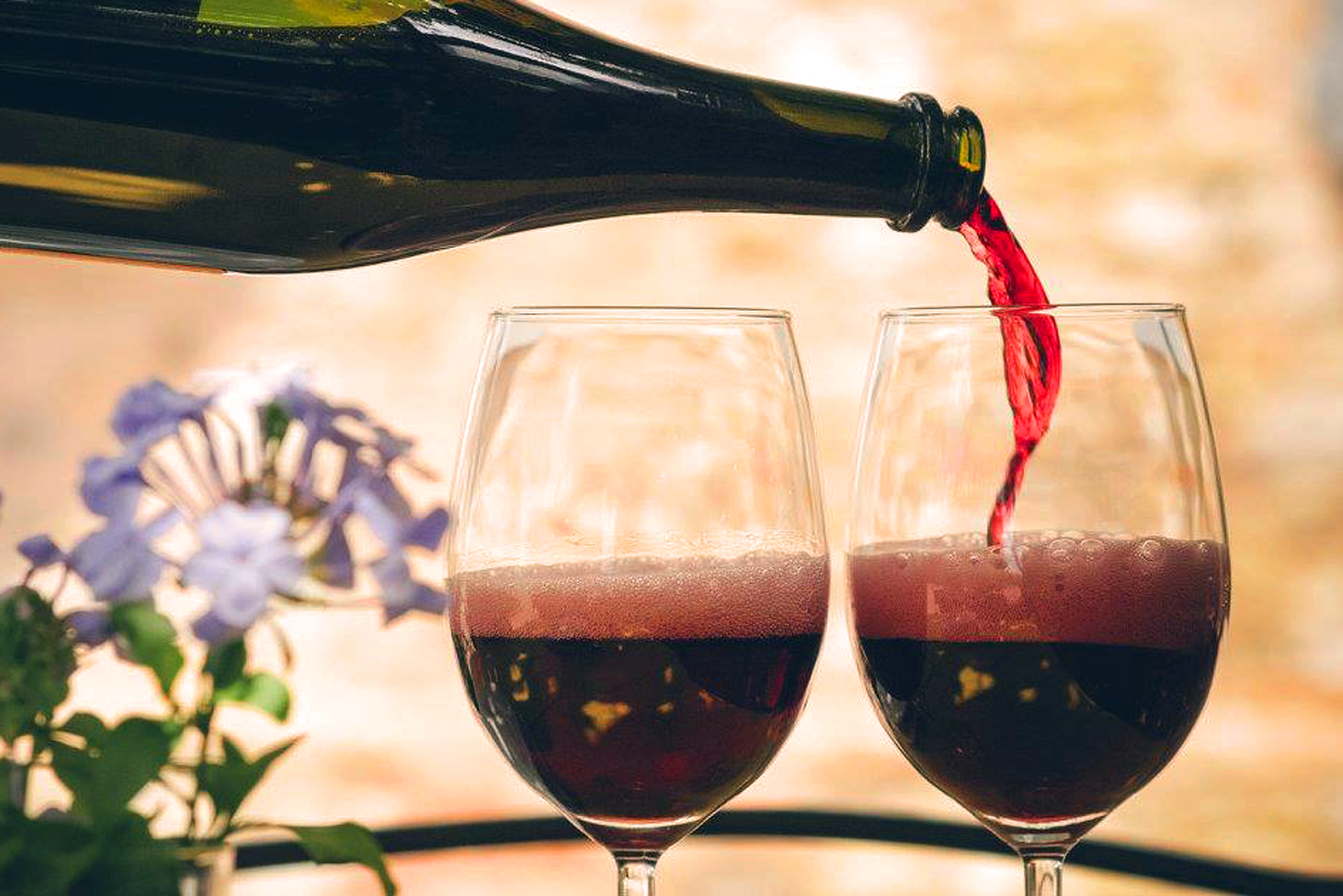 Eine Flasche Lambrusco, aus der perlender roter Schaumwein in zwei Weingläser gegossen wird. Im Hintergrund sanfte, mediterrane Farben und violette Blüten – eine Hommage an den ikonischen italienischen Lambrusco.