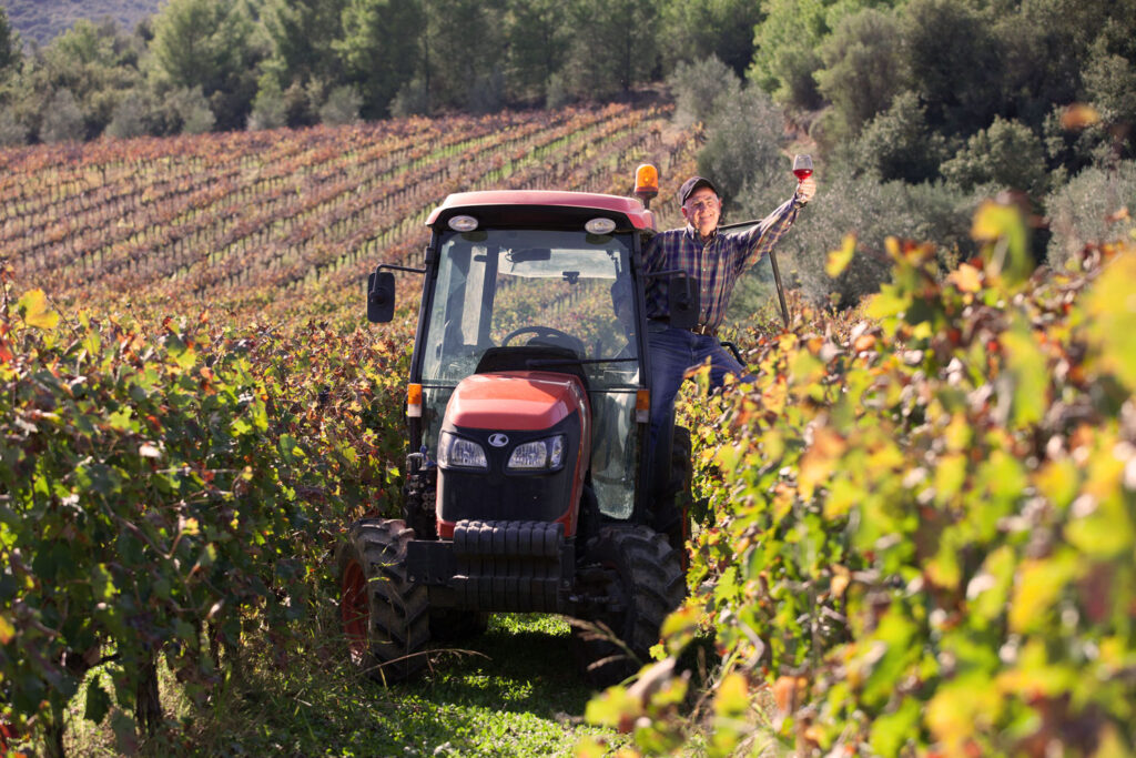Winzer auf einem Traktor mitten in den herbstlichen Lambrusco-Weinbergen bei der Arbeit, mit einem Glas Rotwein in der Hand und einem Lächeln im Gesicht.