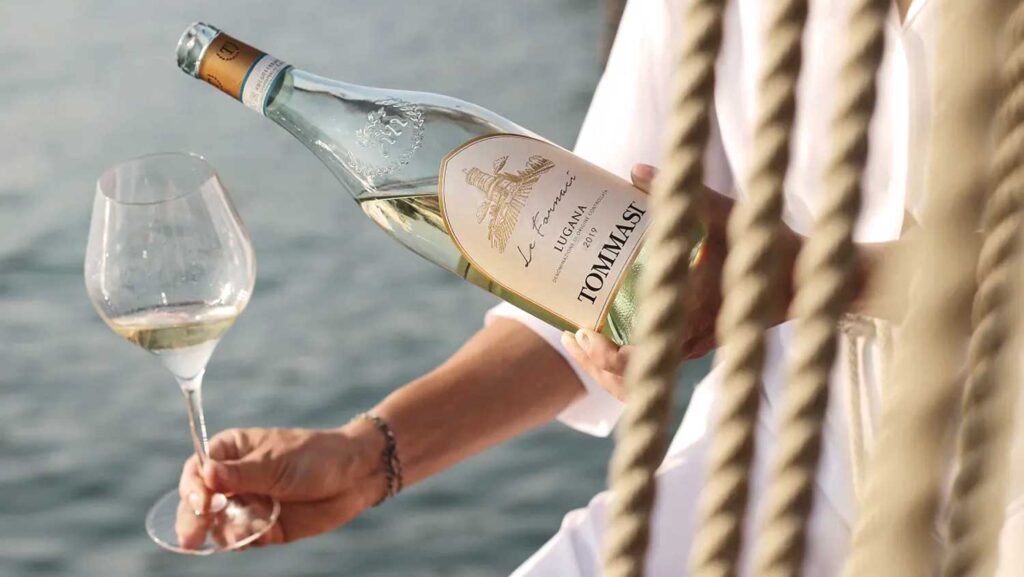 Eine Person gießt ein Glas Lugana-Wein auf einem Boot am Gardasee ein, mit einer stilvollen Weinflasche in der Hand – ein Symbol für italienische Eleganz und Genuss.