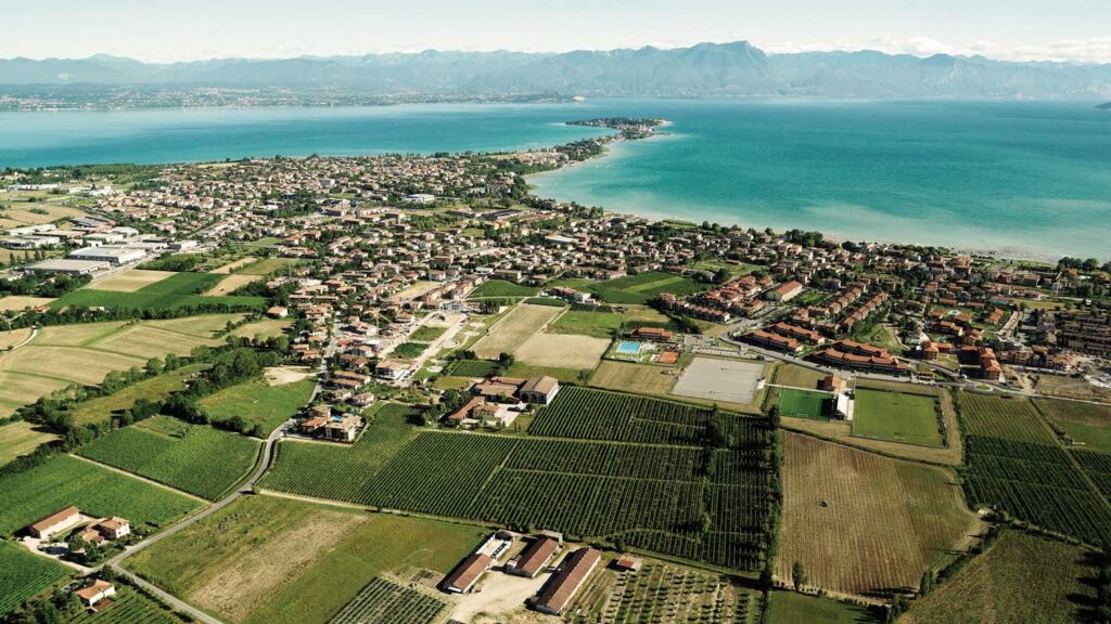 Luftaufnahme eines Küstenortes am Gardasee, umgeben von Weinbergen, die die fruchtbare Landschaft der Lugana-Weinregion zeigen.