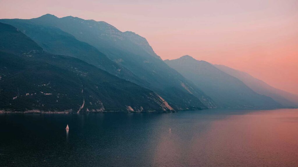 Ein idyllischer Blick auf den Gardasee bei Sonnenuntergang, mit sanftem rosa-orangefarbenem Himmel, ruhigem Wasser und den imposanten Bergen im Hintergrund. Ein kleines Segelboot gleitet über die Wasseroberfläche und verstärkt die Atmosphäre von Ruhe und Naturverbundenheit.