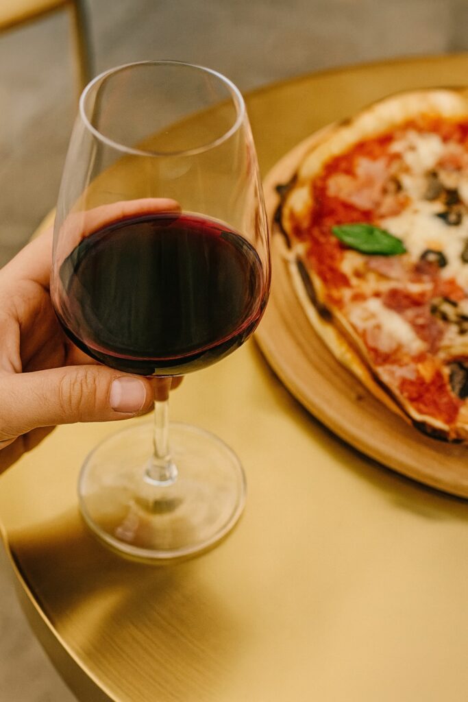 Nahaufnahme eines Glases Rotwein auf einem goldenen Tisch neben einer frisch gebackenen Pizza Margherita. Die warme Beleuchtung hebt die satten Farben des Weins und der Pizza hervor und schafft eine elegante, mediterrane Atmosphäre – ideal für italienische Genussmomente.