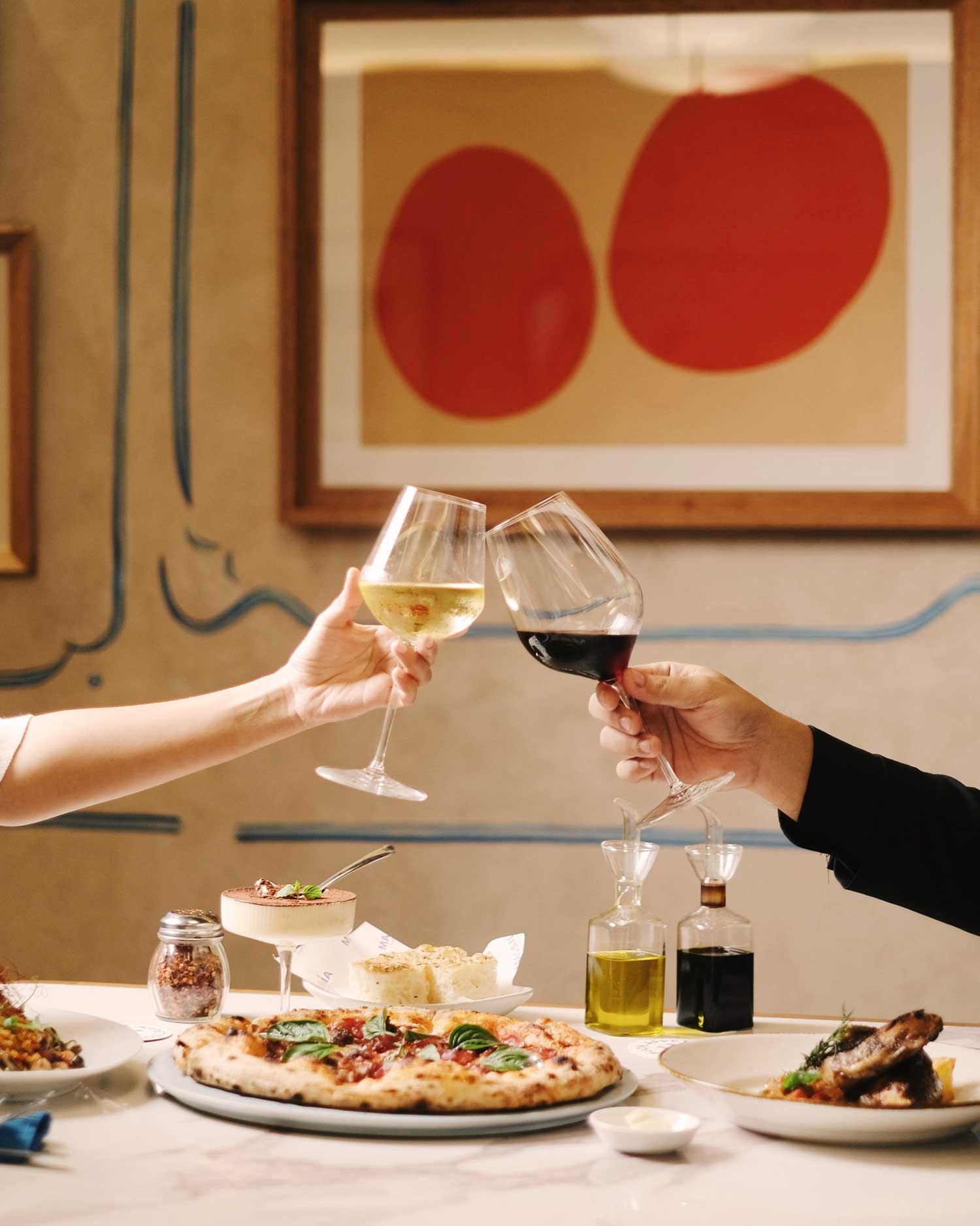 Ein stilvoll gedeckter Tisch in einem italienischen Restaurant, auf dem eine klassische Pizza mit Basilikum serviert wird. Zwei Personen stoßen mit einem Glas Rot- und Weißwein an – ein eleganter Moment mediterraner Lebensfreude.