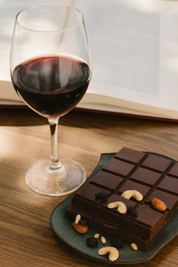 Ein Glas Rotwein steht auf einem glatten Holztisch neben einer Servierplatte mit dunkler Schokolade, garniert mit Cashewnüssen, Mandeln, Pinienkernen und getrockneten Beeren. Im Hintergrund liegt ein geöffnetes Buch, leicht unscharf, in warmem Tageslicht.