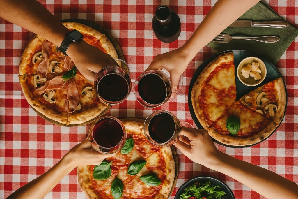 Vier Freunde stoßen mit Gläsern Primitivo Rotwein über einem rustikalen Tisch an, auf dem mehrere klassische italienische Pizzen stehen – Margherita, Prosciutto und Funghi – serviert auf einer rot-weiß karierten Trattoria-Tischdecke. Gesellige italienische Atmosphäre mit frischen Zutaten und mediterranem Flair.