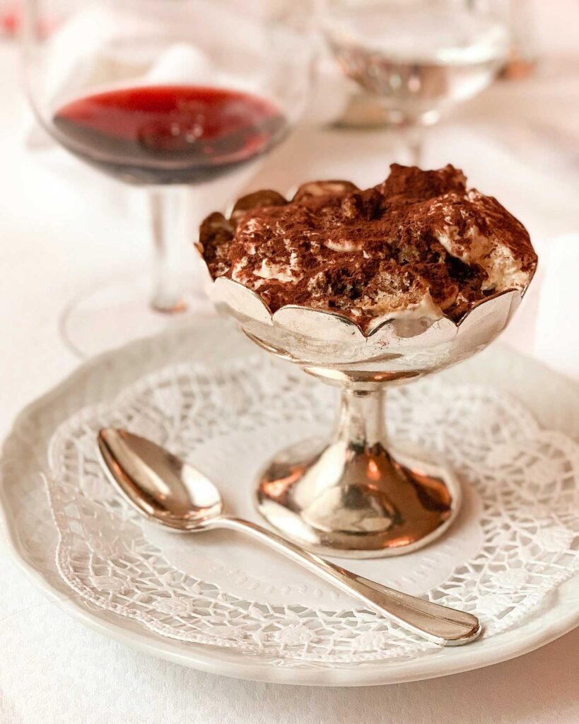 Ein klassisches Tiramisù in einer eleganten silbernen Dessertschale, serviert auf einem weißen Teller mit Spitzenunterlage. Im Hintergrund steht ein Glas Rotwein – die perfekte italienische Nachspeise.