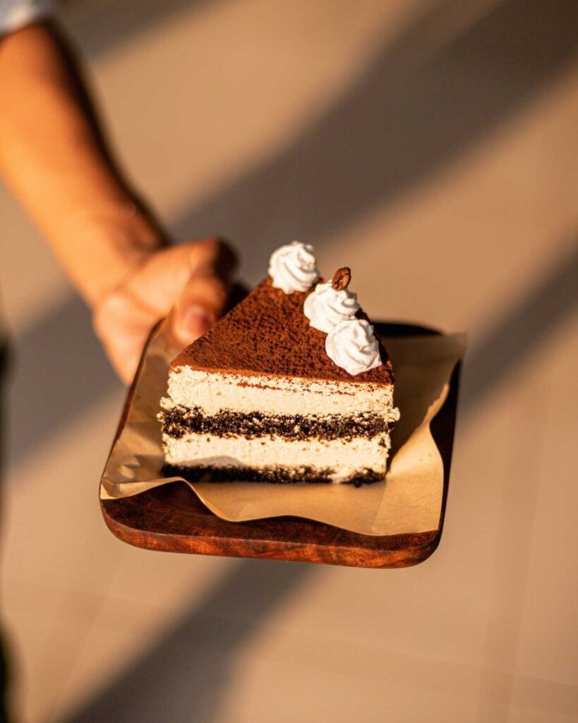 Ein köstliches Stück Tiramisù auf einem Holztablett, mit dunklem Kakaopulver bestäubt und mit kleinen Sahnetupfern verziert. Der warme Lichteinfall betont die cremige Konsistenz des Desserts.