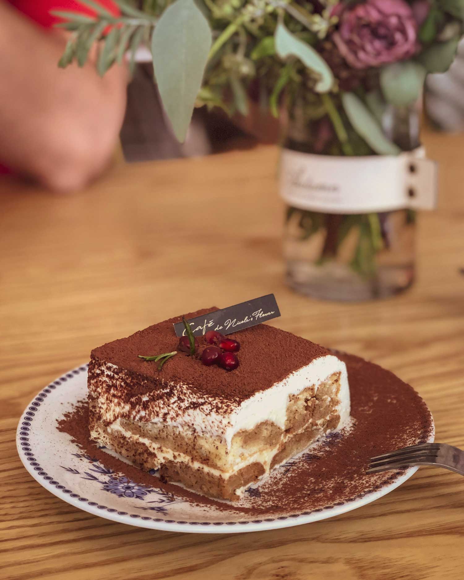 Ein kunstvoll serviertes Tiramisù auf einem antiken Porzellanteller, mit dekorativen Kakaopulver-Schichten und einer kleinen Beerenverzierung. Ein wahrer Hingucker auf dem Holztisch, begleitet von einem Blumenarrangement.