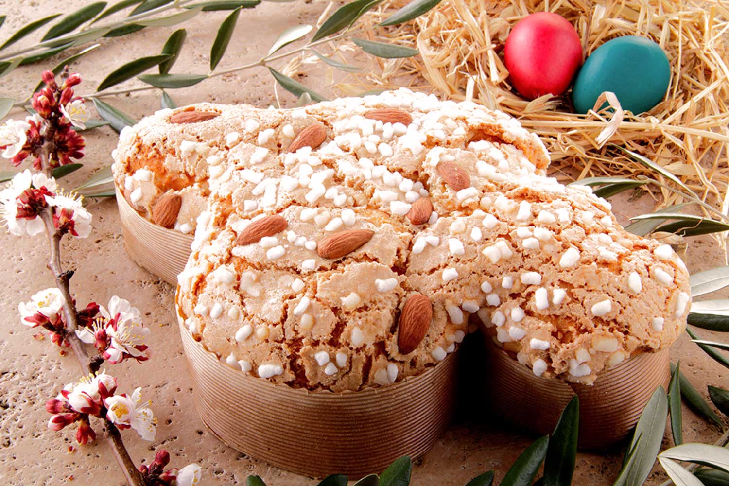 Traditionelle italienische Colomba Pasquale, ein Ostergebäck in Taubenform, mit knuspriger Mandel- und Zuckerglasur. Im Hintergrund dekorative Olivenzweige und bunte Ostereier auf Stroh