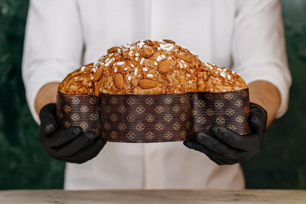 Konditor präsentiert eine kunstvoll gebackene Colomba Pasquale mit dunkler Papierform, goldener Kruste und Mandeln – italienische Backkunst zu Ostern.