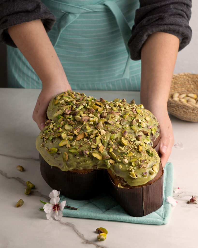 Colomba Pasquale mit Pistazienglasur und gehackten Pistazien, gehalten von einer Person in Schürze – kreative Oster-Variation mit sizilianischer Note.