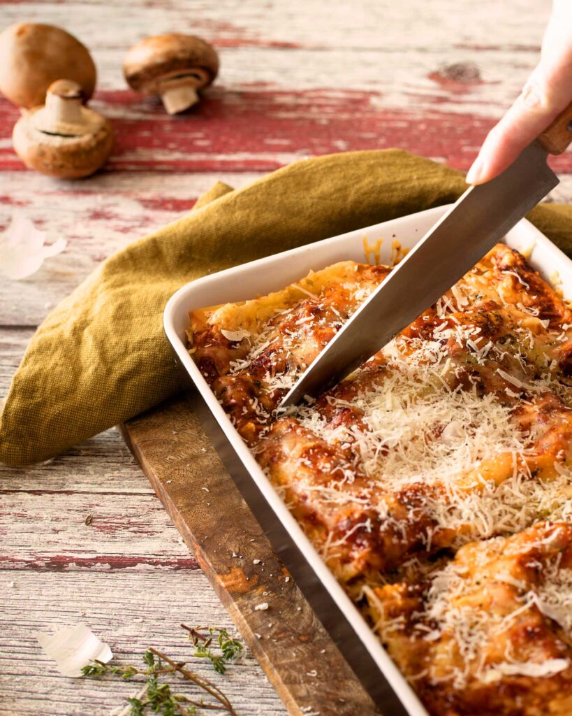 Eine Lasagne al Forno wird mit einem großen Messer in Stücke geschnitten, während Parmesan darüber gerieben wird. Im Hintergrund sind Champignons zu sehen.