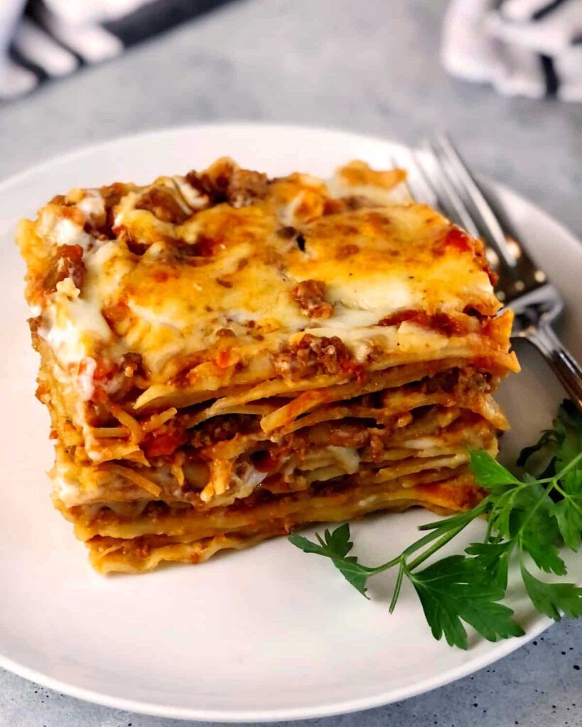 Ein Stück Lasagne al Forno auf einem weißen Teller, geschichtet mit Bolognese, Bechamel und geschmolzenem Käse, garniert mit Petersilie.