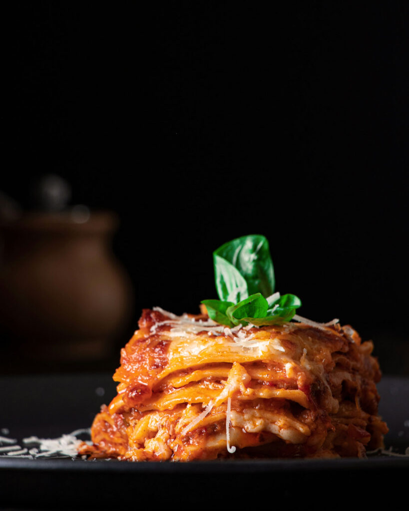 Lasagne al forno mit Tomatensugo und Béchamel, kunstvoll geschichtet und mit frischem Basilikum garniert – italienisches Soulfood in Nahaufnahme auf schwarzem Teller vor dunklem Hintergrund.