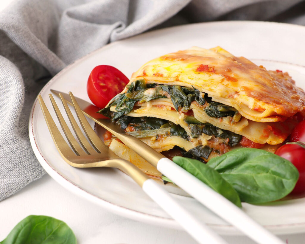 Vegetarische Lasagne mit Spinat und Tomatensauce auf weißem Teller, serviert mit Kirschtomaten, Basilikumblättern und goldfarbenem Besteck – eine leichte Variante des Klassikers.