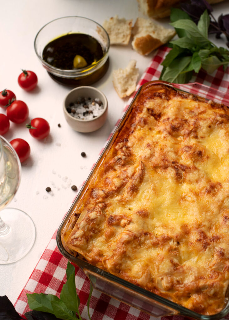 Lasagne frisch aus dem Ofen in einer Glasform auf rot-weiß karierter Tischdecke, umgeben von Kirschtomaten, Olivenöl, Brot und Gewürzen – rustikaler Genuss wie bei der Nonna.