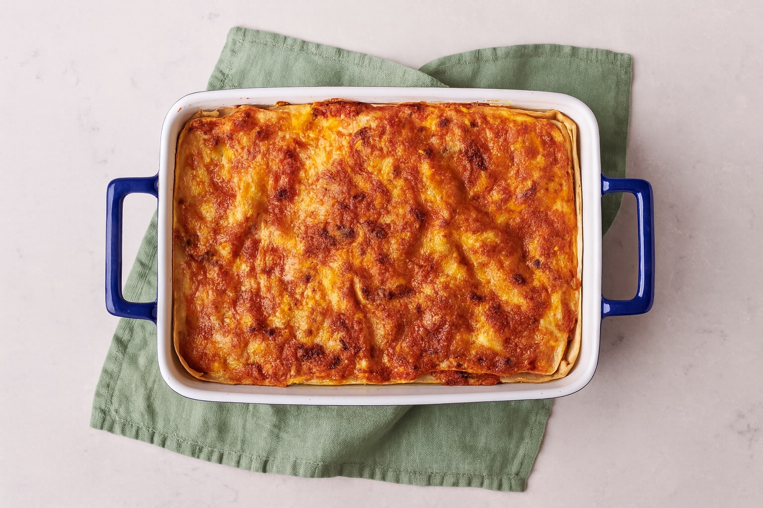 Überbackene Lasagne in einer weißen Ofenform mit blauen Griffen, goldbraune Käsekruste, serviert auf grünem Leinentuch – minimalistisches Stillleben mit mediterranem Charakter.
