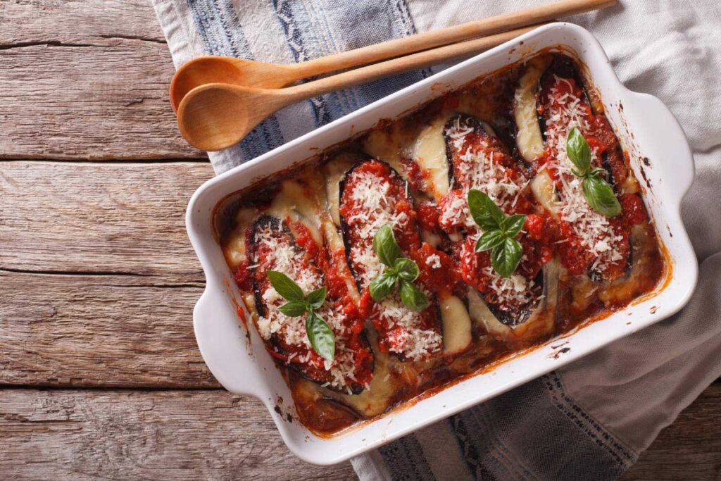 Ofenfrische Parmigiana di Melanzane in einer weißen Auflaufform, belegt mit geschmolzenem Käse, Tomatensauce und frischen Basilikumblättern