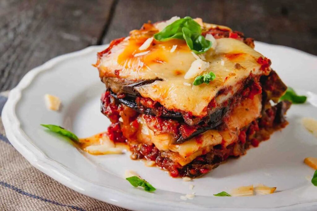 Ein Stück geschichtete Parmigiana di Melanzane auf einem weißen Teller, mit geschmolzenem Käse, frischen Tomaten und Basilikumblättern dekoriert