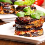 Mini-Portionen von Parmigiana di Melanzane, kunstvoll geschichtet mit Auberginen, Tomatensauce und Mozzarella, serviert auf weißen Tellern mit frischen Basilikumblättern