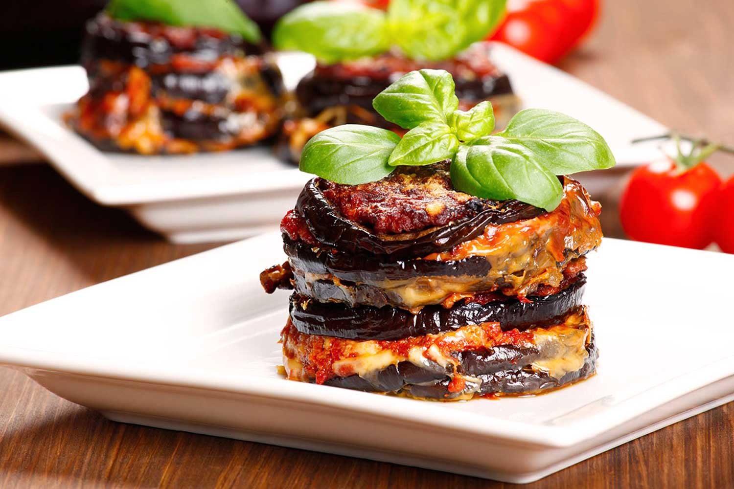 Mini-Portionen von Parmigiana di Melanzane, kunstvoll geschichtet mit Auberginen, Tomatensauce und Mozzarella, serviert auf weißen Tellern mit frischen Basilikumblättern