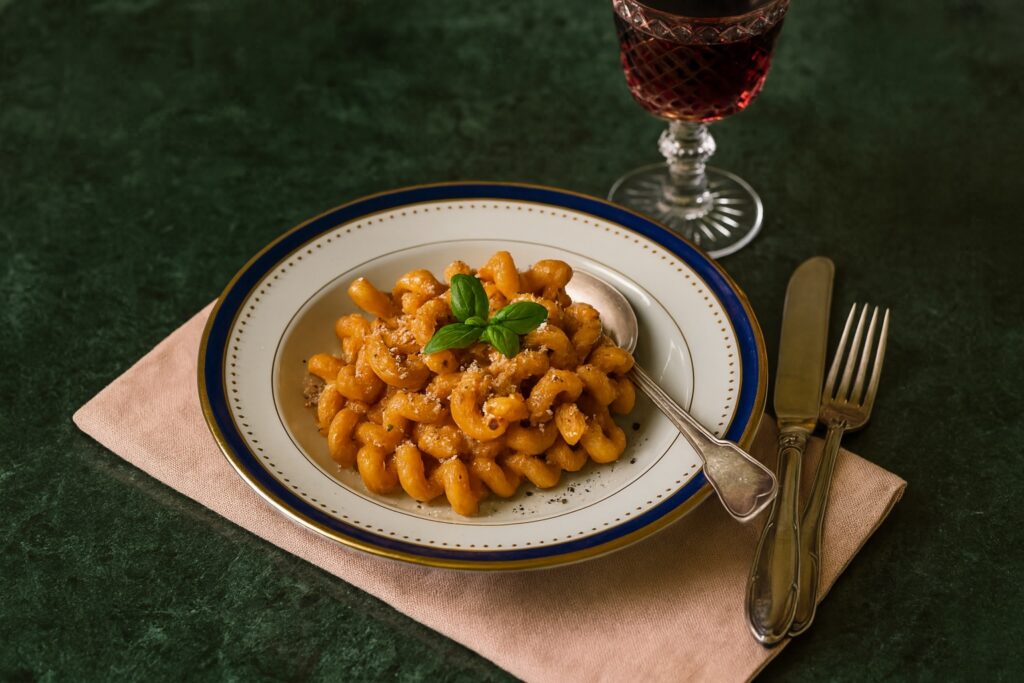 Teller mit gedrehten Pasta in cremigem Pesto Rosso, garniert mit frischem Basilikum, serviert auf edlem Porzellan mit Besteck und einem Glas Rotwein.