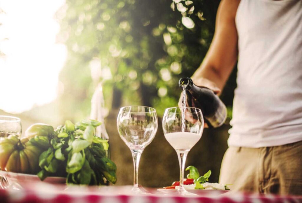 Ein Mann in sommerlicher Kleidung schenkt draußen an einem gedeckten Tisch ein Glas Prosecco ein, umgeben von frischen Zutaten wie Basilikum und Tomaten, mit einer unscharfen, grünen Gartenkulisse im Hintergrund.