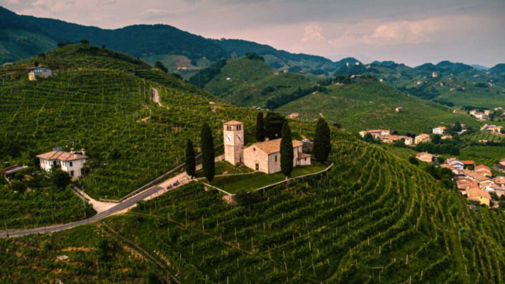 Eine historische Kirche auf einem Hügel umgeben von grünen Prosecco-Weinbergen in der Region Veneto. Die malerische Landschaft steht für das Herzstück der Prosecco-Produktion.