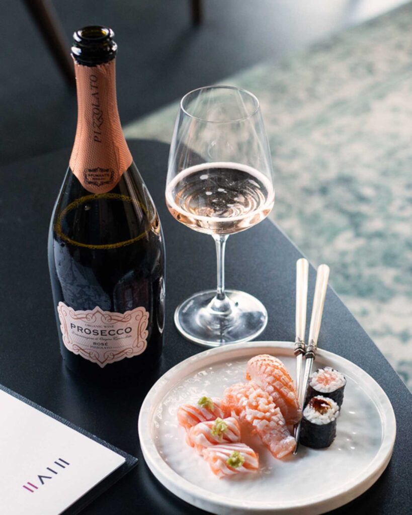 Eine geöffnete Flasche Prosecco Rosé neben einem gefüllten Weinglas, begleitet von einem Teller mit Sushi, darunter Lachs-Nigiri und Maki-Rollen. Die elegante Präsentation zeigt die perfekte Kombination von Prosecco Rosé mit feiner japanischer Küche.