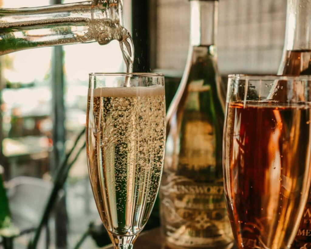 Ein Glas Prosecco wird frisch aus einer Flasche eingeschenkt, wobei feine Perlen aufsteigen. Im Hintergrund stehen weitere Flaschen mit italienischem Schaumwein sowie ein Glas mit Rosé-Prosecco. Die Szene vermittelt eine elegante Atmosphäre, perfekt für einen stilvollen Aperitivo.