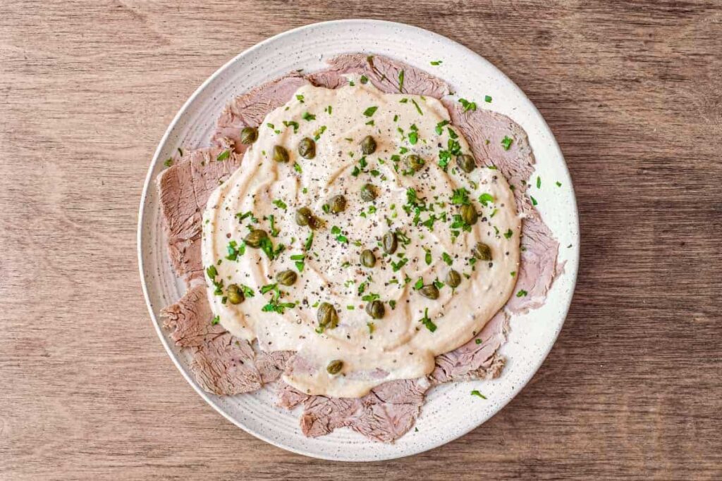 Klassisches Vitello Tonnato auf einem weißen Teller – dünne Kalbfleischscheiben, großzügig mit Thunfischsauce bedeckt und mit Kapern sowie Petersilie bestreut.