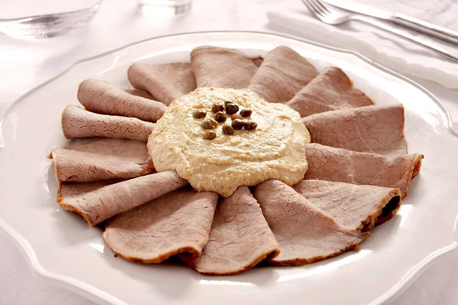 Elegante Präsentation von Vitello Tonnato mit dünnen Kalbfleischscheiben, die fächerförmig um eine Thunfisch-Kapern-Sauce arrangiert sind, serviert auf einem weißen Porzellanteller.