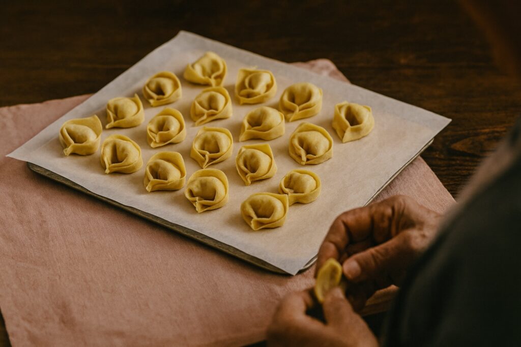 Auf einem mit Backpapier belegten Blech ruhen gleichmäßig geformte Tortellini, bereit zum Kochen – Symbol für Geduld und Perfektion.