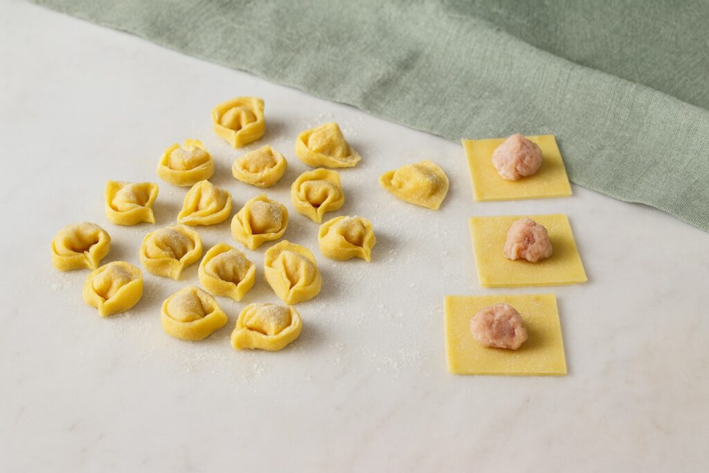 Frisch gefüllte Teigquadrate mit Fleischfüllung und fertige Tortellini nebeneinander auf einer hellen Arbeitsfläche mit grünem Tuch.