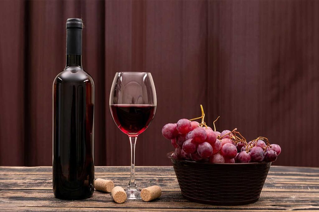 Eine Flasche Rotwein neben einem Glas mit eingeschenktem Wein und einer Schale roter Trauben auf einem rustikalen Holztisch.