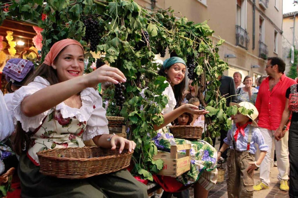 Junge Frauen in traditioneller Kleidung feiern das Weinfest und verteilen Trauben an Kinder und Zuschauer.