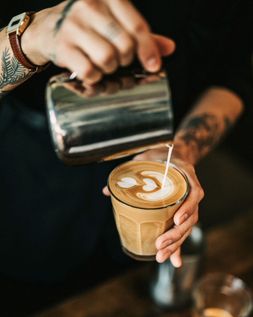 Ein Barista gießt kunstvoll Milchschaum in ein Glas Cappuccino und zaubert ein Herzmotiv auf den cremigen Milchschaum.