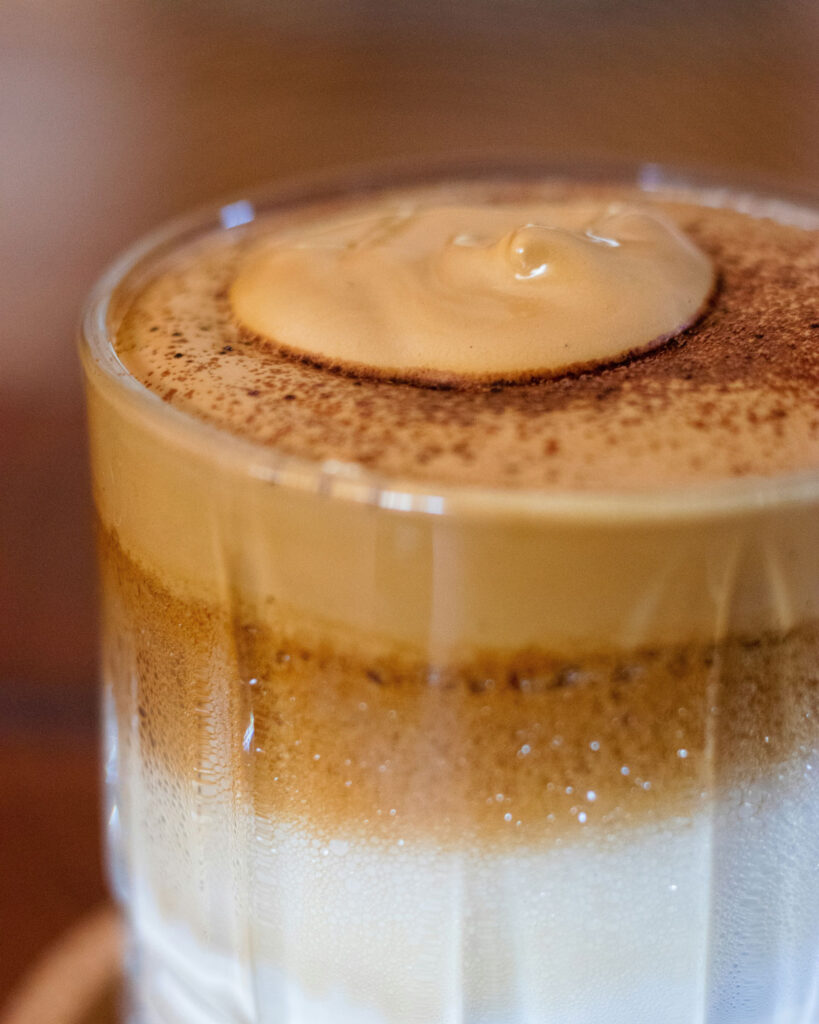 Nahaufnahme eines geschichteten Cappuccino mit dickem Schaum und einer Prise Kakao, die dem Getränk den letzten Schliff verleiht.