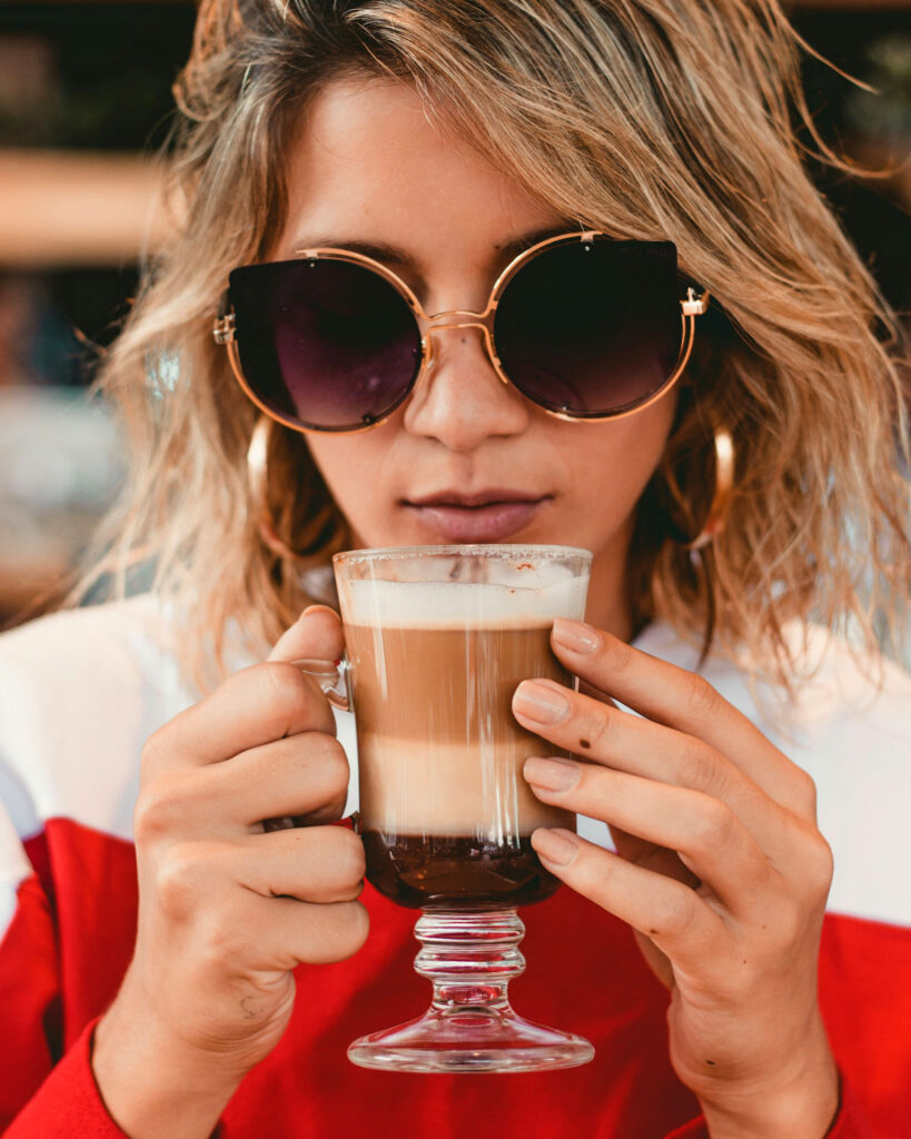 Junge Frau mit großen Sonnenbrillen und roten Pullover hält ein elegantes Glas mit einem perfekt geschichteten Cappuccino in den Händen und genießt den Moment.